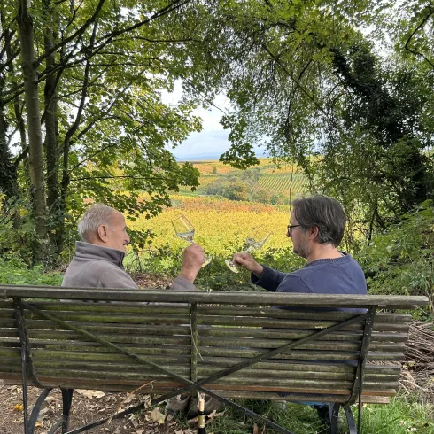 Zwei Männer sitzen auf einer Bank und stoßen mit Gläsern an. Im Hintergrund sieht man eine schöne, grüne Landschaft mit Weinreben.