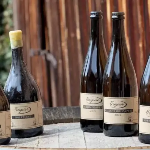 Eine Auswahl von fünf Flaschen Schaumwein steht auf einem Holzfass. Die Flaschen haben unterschiedliche Etiketten und Größen.