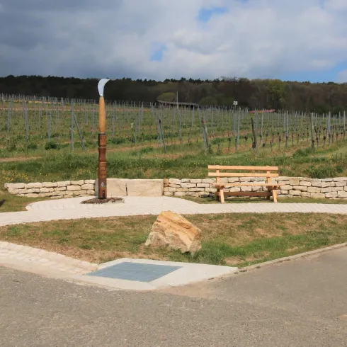 Ein kleiner Platz inmitten von Weinbergen mit einer Bank aus Holz. Im Hintergrund sind die sanften Hügel der Landschaft zu sehen.