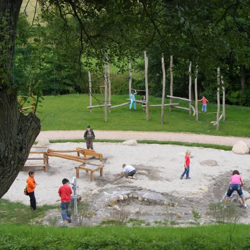 Ein Spielplatz in der Natur mit Kindern, die sich im Sand austoben. Im Hintergrund sind Klettergerüste und Grünflächen sichtbar.