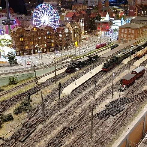 Ein detailliertes Modellbahn-Szenario mit mehreren Gleisen und Zügen. Im Hintergrund sind beleuchtete Gebäude und ein Riesenrad zu sehen.