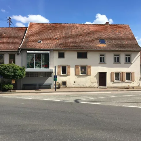 Ein älteres Gebäude mit einem roten Dach und Fensterläden. Es steht an einer Straßenecke, umgeben von weiteren Häusern und Bäumen.