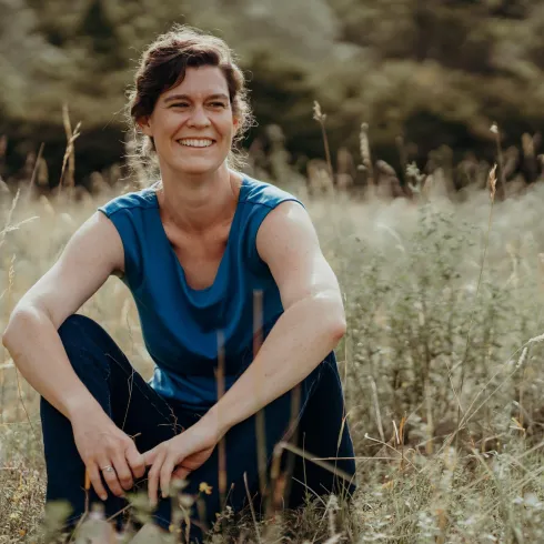 Eine lächelnde Frau sitzt im Gras und genießt die Natur. Sie trägt ein blaues T-Shirt und wirkt entspannt.