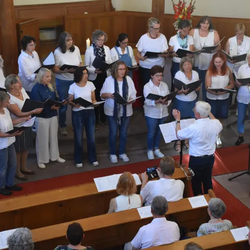 Ein gemischter Chor singt in einer Kirche. Der Chorleiter steht vorne und leitet die Sänger.