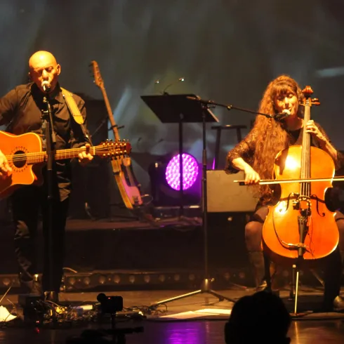 Ein Musiker singt und spielt Gitarre, während eine Cellistin neben ihm spielt. Hintergrundlich sind bunte Lichter zu sehen, die eine lebendige Atmosphäre schaffen.