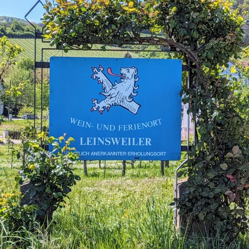 Ein Schild zeigt den Ort Leinsweiler als Wein- und Ferienort an. Umgeben von üppigem Grün und Weinbergen ist es ein malerischer Anziehungspunkt.
