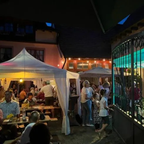Eine lebhafte Sommernacht mit vielen Menschen, die in einem gemütlichen Garten feiern. Zelte und stimmungsvolle Beleuchtung sorgen für eine einladende Atmosphäre.
