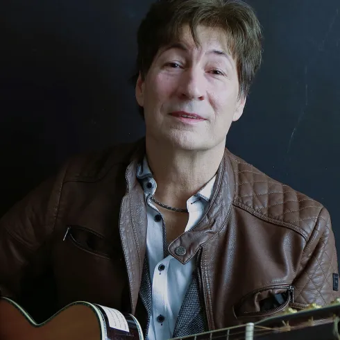 Ein Mann mit einer Gitarre lächelt in die Kamera. Er trägt eine Lederjacke und sitzt vor einem dunklen Hintergrund.