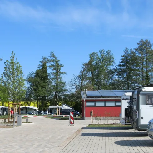 Ein ruhiger Campingplatz mit Wohnmobilen und einem modernen Servicegebäude. Umgeben von Bäumen und einem klaren blauen Himmel.