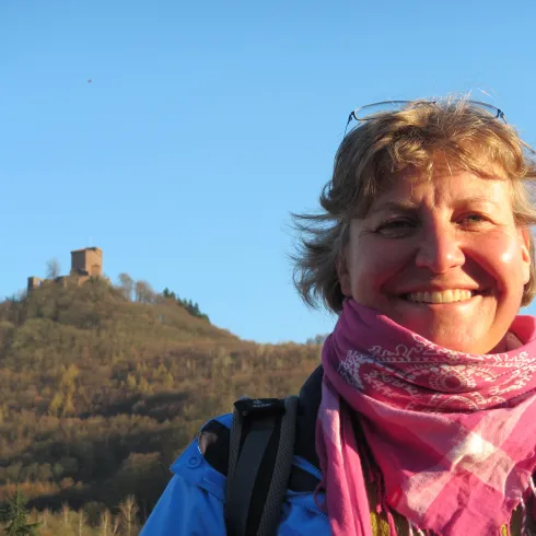 Eine lächelnde Frau steht vor einem hügeligen Landschaftshintergrund mit einer Burgruine. Der Himmel ist klar und blau.