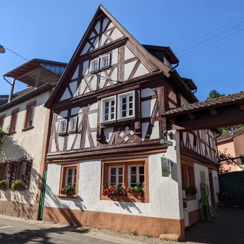 Ein charmantes Fachwerkhaus mit bunten Blumen in den Fenstern. Die Straße ist ruhig und von Sonnenschein beleuchtet.