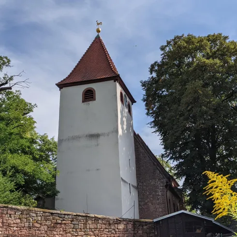 Ein historischer Kirchturm mit rotem Dach, umgeben von hohen Bäumen. Die Szene strahlt eine ruhige und ländliche Atmosphäre aus.