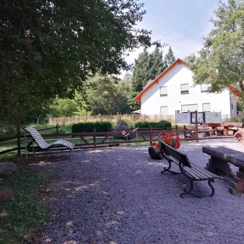 Ein gemütlicher Außenbereich mit Bänken und einem Tisch aus Holz. Im Hintergrund ist ein helles Haus und viel Grün zu sehen.