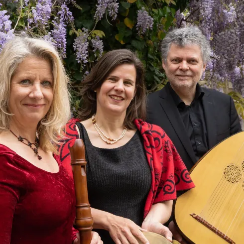 Drei Musiker stehen vor einer Wand mit lila blühenden Pflanzen. Sie halten verschiedene Musikinstrumente und lächeln in die Kamera.