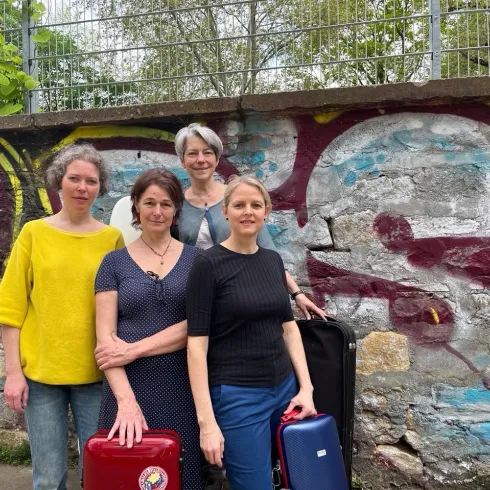 Vier Frauen stehen vor einer bunten Graffiti-Wand. Sie halten Koffer und blicken lächelnd in die Kamera.
