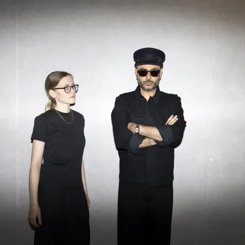 Ein Musiker singt und spielt Gitarre, während eine Cellistin neben ihm spielt. Hintergrundlich sind bunte Lichter zu sehen, die eine lebendige Atmosphäre schaffen.