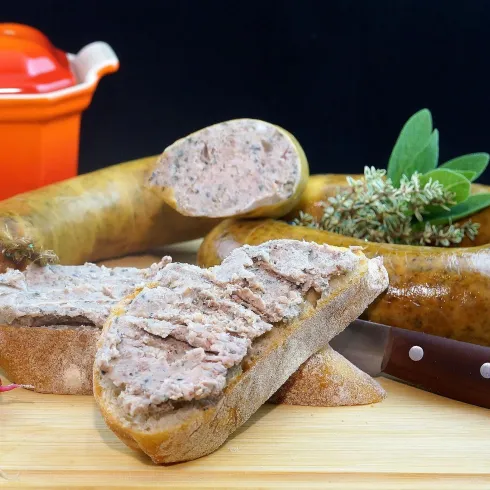 Ein Teller mit geschnittener Wurst, umgeben von frischen Kräutern und Radieschen. Im Hintergrund ist ein roter Topf und ein Küchenmesser sichtbar.