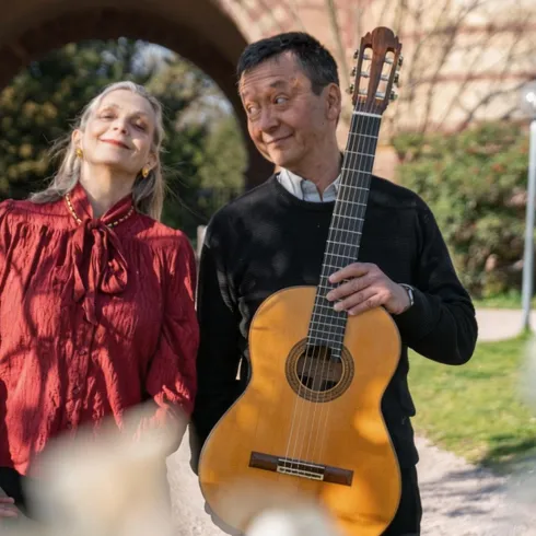 Ein Mann hält eine Gitarre und lächelt, während eine Frau neben ihm steht. Im Hintergrund sind Blumen und ein schöner Park zu sehen.
