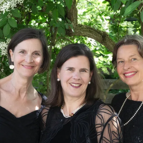 Drei Frauen in eleganten schwarzen Kleidern stehen lächelnd vor einem Hintergrund mit blühenden Pflanzen. Sie scheinen einen festlichen Anlass zu genießen.