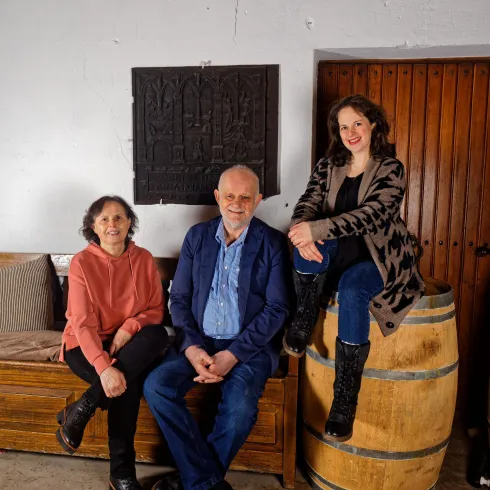 Drei Personen sitzen in einer rustikalen Umgebung. Sie lächeln und scheinen entspannt zu sein.