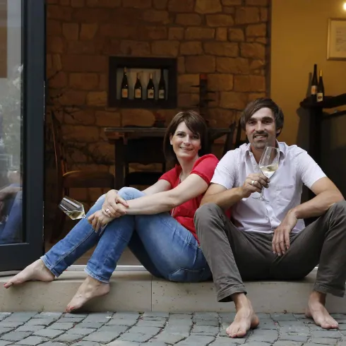 Ein Paar sitzt entspannt auf den Stufen vor einem Haus und genießt ein Getränk. Im Hintergrund sind Weinflaschen und eine gemütliche Inneneinrichtung zu sehen.