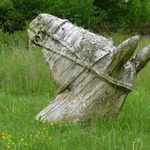 Eine Holzskulptur in Form eines Pferdes steht in einer grünen Wiese. Umgeben von Bäumen und Wiesenblumen wirkt sie rustikal und natürlich.
