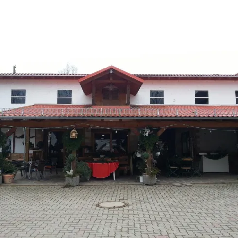 Ein traditionelles Gebäude mit einem roten Dach und einer überdachten Terrasse. Vor dem Eingang stehen dekorative Pflanzen und eine einladende Atmosphäre.