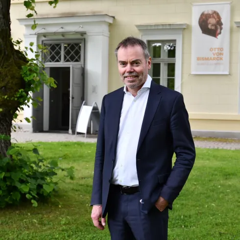 Ein Mann steht in einem grünen Garten vor einem historischen Gebäude. Im Hintergrund befindet sich ein großes Fenster und ein Plakat.