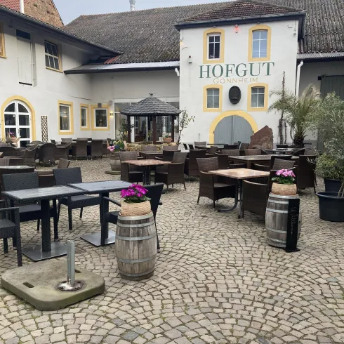 Ein gemütlicher Außenbereich eines Restaurants mit Tischen und Stühlen. Im Hintergrund befindet sich ein traditionelles Gebäude mit der Aufschrift "HOFGUT".