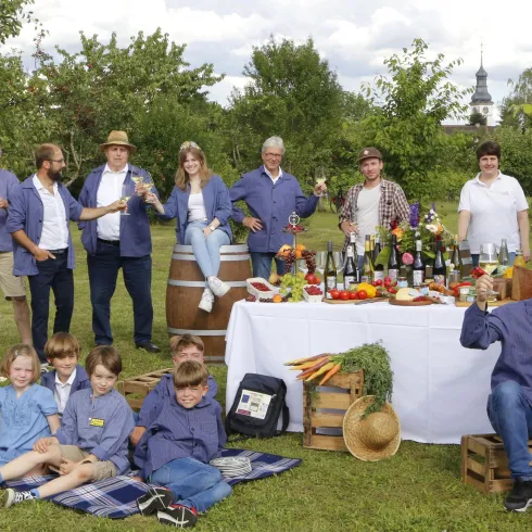 Eine Gruppe von Menschen feiert im Freien mit Essen und Getränken. Im Hintergrund stehen Obstbäume und ein schön gedeckter Tisch.