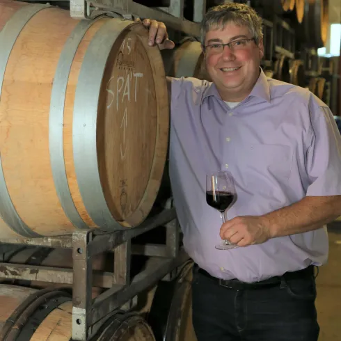 Ein Mann steht neben Weinfässern in einem Weinkeller und hält ein Glas Rotwein in der Hand. Er trägt ein lila Hemd und lächelt freundlich.