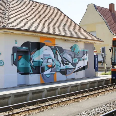 Eine Straßenbahn hält an einer Station mit einem bunten Graffiti an der Wand. Die Umgebung ist hell und einladend.