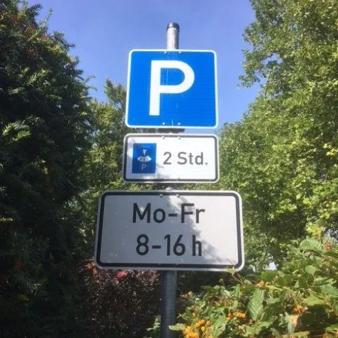 Ein Parkplatzschild mit der Aufschrift „2 Std.“ und den Öffnungszeiten „Mo-Fr 8-16 h“. Umgeben von Bäumen und Büschen.