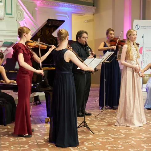 Eine Musikgruppe führt ein Konzert auf einer eleganten Bühne auf. Die Musikerinnen spielen verschiedene Instrumente und tragen festliche Kleider.