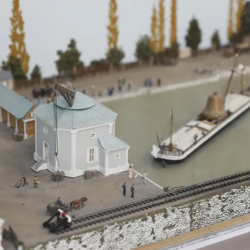 Eine detaillierte Modellansicht eines Hafenbereichs mit einem Schiff und einer kleinen Hafenanlage. Umgeben ist die Szene von Bäumen und Figuren, die den Alltag im Hafen darstellen.