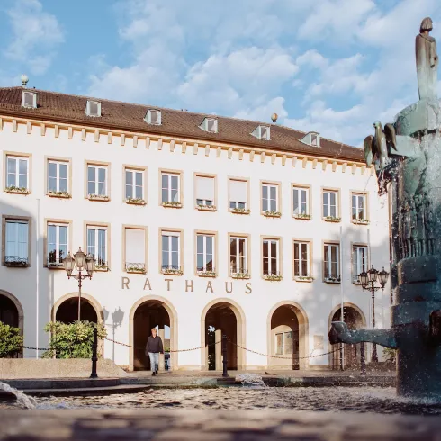 Ein historisches Rathaus mit mehreren Fenstern und einer großen Eingangstür. Im Vordergrund steht ein Brunnen mit skulpturalen Elementen.