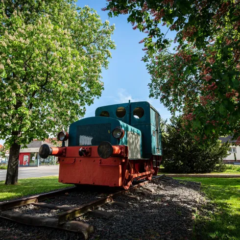 Eine alte Lokomotive steht auf Schienen, umgeben von grünen Bäumen. Im Hintergrund sind einige Gebäude sichtbar.