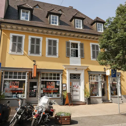 Ein Farbiges Gebäude mit gelber Fassade und mehreren Fenstern. Davor stehen Motorräder und ein paar Bäume.