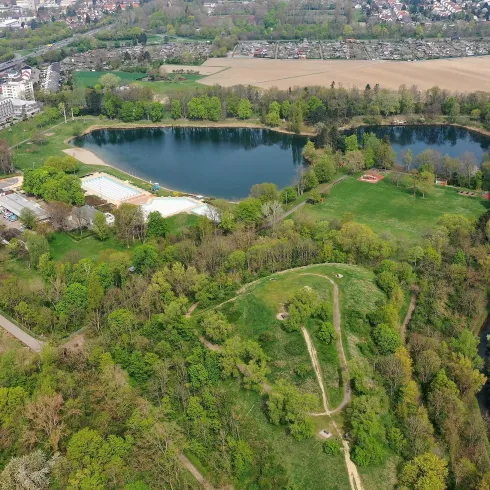 Eine grüne Landschaft mit einem See und viel Vegetation. Im Hintergrund sind Felder und städtische Gebäude zu sehen.