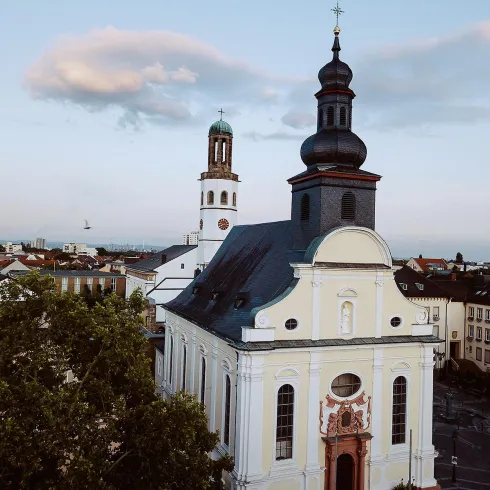 Eine schöne Kirche mit einer markanten Kuppel und einem hohen Turm. Im Hintergrund sind einige Gebäude und ein klarer Himmel zu sehen.