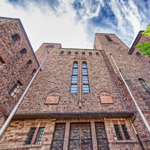 Ein historisches Gebäude aus Stein mit hohen Türmen und großen Fenstern. Die Szenerie wird von einem teilweise bewölkten Himmel und dem Grün von Bäumen ergänzt.