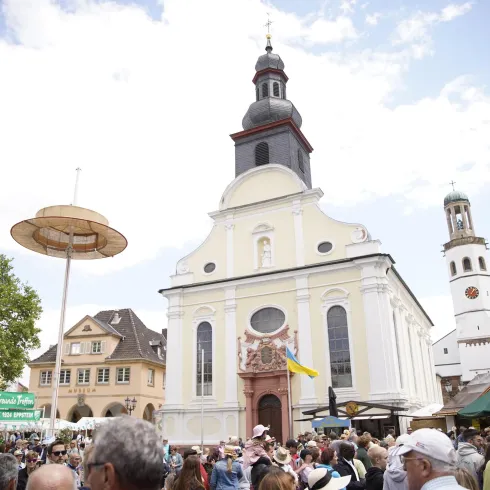 Ein belebter Marktplatz mit einer großen Kirche im Hintergrund. Menschen stehen zusammen und genießen die Atmosphäre.