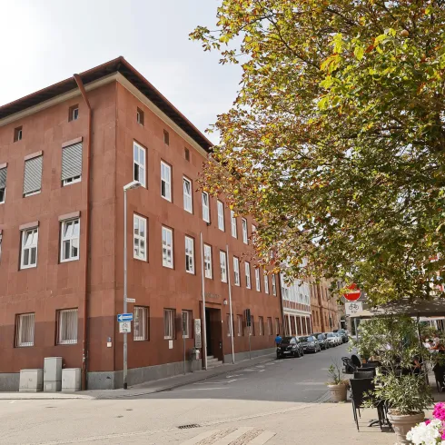 Ein rotes Gebäude an einer Straßenecke, umgeben von Bäumen und Pflanzen. Die Straße wirkt ruhig und urban.