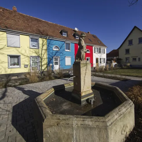 Ein schöner Platz mit bunten Häusern und einem Brunnen in der Mitte. Die Architektur ist modern und die Umgebung ist gepflegt.