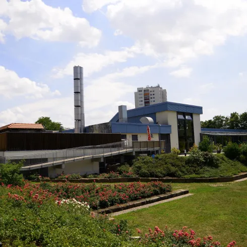 Ein modernes Gebäude mit einem blauen Dach und vielen Fenstern. Im Vordergrund befindet sich ein schön angelegter Garten mit Blumen und Bäumen.