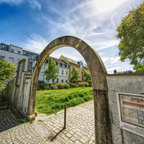 Ein schöner Bogen aus Stein führt in einen grünen Garten. Im Hintergrund sind Häuser und blauer Himmel zu sehen.