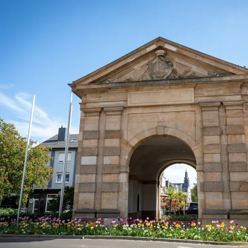 Ein historisches Tor mit einer dekorativen Fassade. Rund um das Tor blühen bunte Blumen und es ist ein klarer blauer Himmel zu sehen.