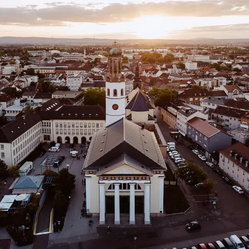 Eine Stadtansicht mit einer Kirche im Vordergrund und einer Wohngegend im Hintergrund. Die Sonne geht am Horizont unter und schafft eine warme Atmosphäre.