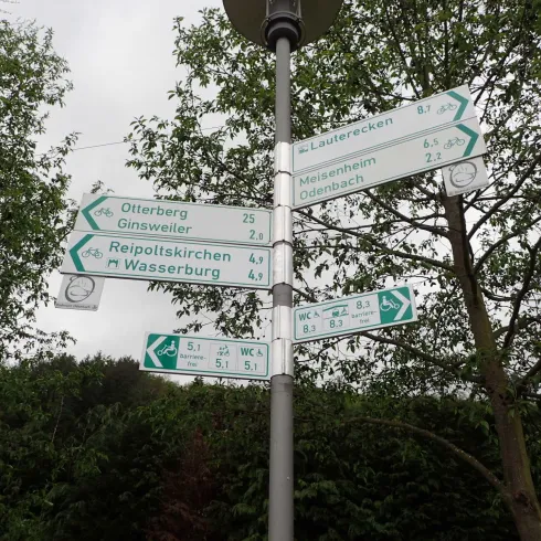 Ein Wegweiser zeigt verschiedene Richtungen mit Entfernungen zu mehreren Orten. Umgeben von Bäumen, gibt es zahlreiche grüne Schilder.