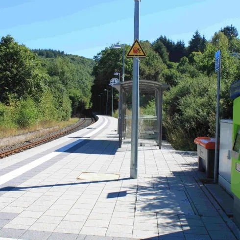 Eine ruhige Bahnhofshaltestelle mit einem Wartenbereich und einer Ticketautomaten. Im Hintergrund sind grüne Bäume und ein samtblauer Himmel zu sehen.